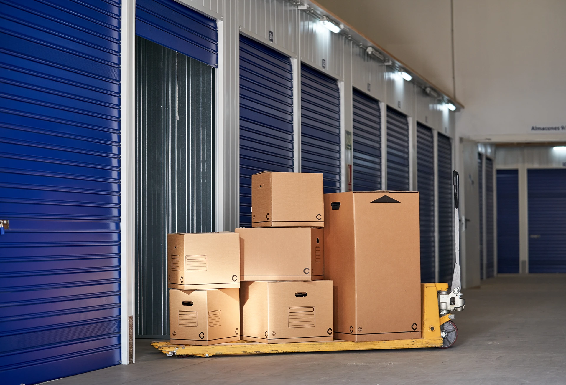 Cardboard boxes on a yellow pallet jack in front of storage units.